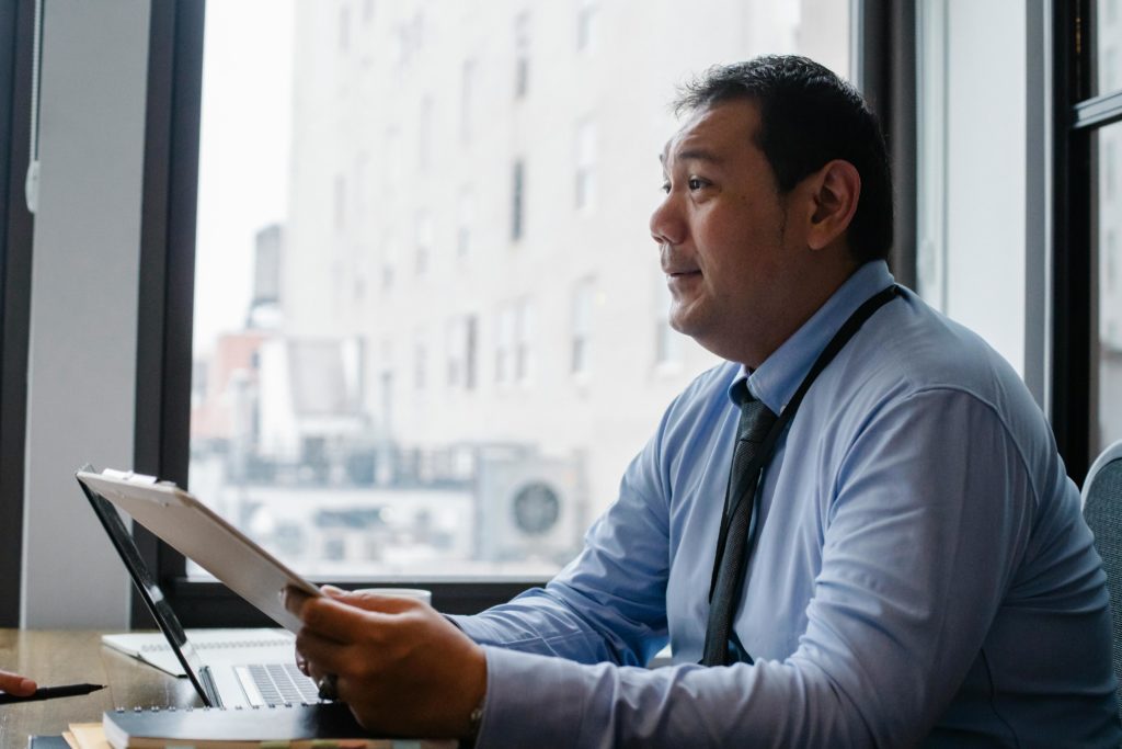 Side view of ethnic male boss with clipboard at table with modern gadget near window
