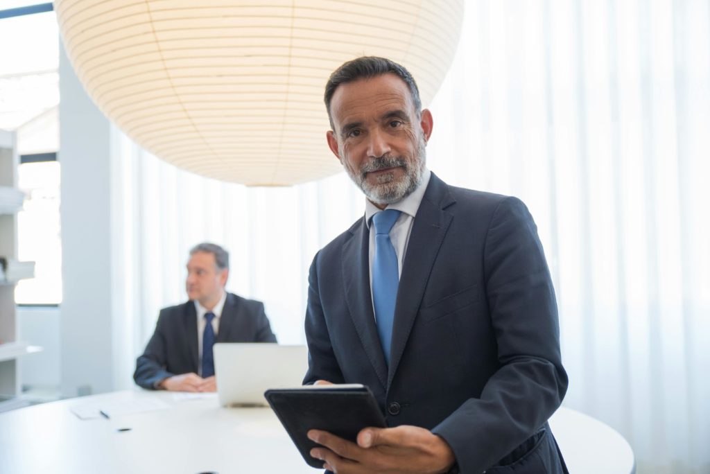 Two businessmen in a modern office, one holding a tablet and the other working on a laptop.